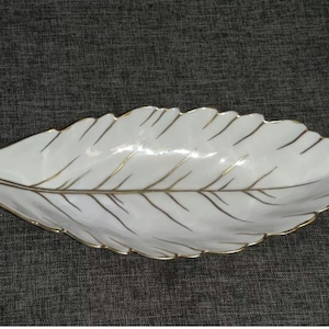 Puede incluir: Un plato de servir de porcelana blanca con forma de hoja con detalles dorados. El plato está diseñado para parecerse a una sola hoja con una vena central y venas más pequeñas que se ramifican.