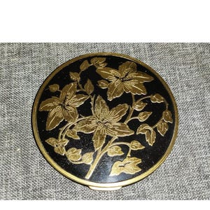 Op de afbeelding: Een ronde, zwarte compact met een gouden bloemenontwerp. De bloemen zijn gestileerd en hebben een delicaat, ingewikkeld ontwerp.