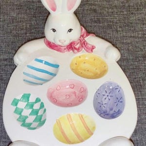 Può includere: Un vassoio da portata a forma di coniglietto pasquale in ceramica bianca con sei incavi a forma di uovo. Il coniglietto ha orecchie foderate di rosa e una sciarpa rosa. Le uova sono dipinte con vari motivi e colori, tra cui strisce blu, quadri verdi e cuori rosa.