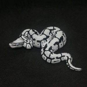 May include: A white and black patterned snake figurine. The snake is coiled and has a detailed design with black markings on a white base. The snake's head is visible, with small blue eyes.
