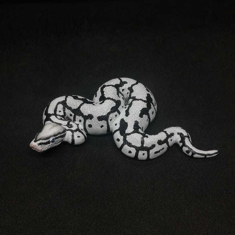 May include: A white and black snake figurine with a detailed pattern. The snake is coiled and has a realistic appearance, with a textured surface and a dark background.