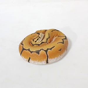 May include: A coiled, ceramic snake figurine. The snake is primarily tan with black markings and a white underbelly. The figurine is a detailed representation of a snake, likely a ball python, and is set against a white background.