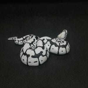May include: A white and black snake figurine, coiled on a dark surface. The snake has a pattern of black blotches and spots on a white background. The head is raised, and the tail is curled.