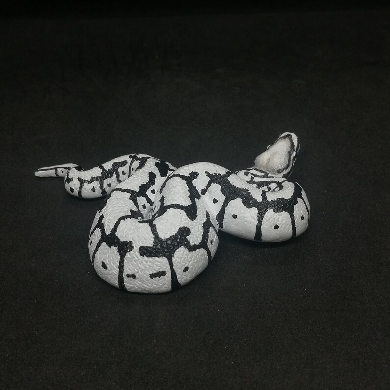 May include: A white and black snake figurine, coiled on a dark surface. The snake has a pattern of black blotches and spots on a white background. The head is raised, and the tail is curled.