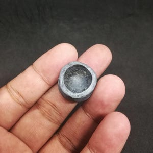 May include: A small, gray, stone-like bowl with a circular shape. The bowl has a rough, textured exterior and a smooth, concave interior. The inside of the bowl appears to have a slightly reflective, glittery surface.