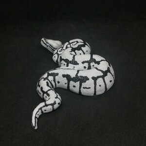 May include: A white and black snake figurine, coiled on a dark surface. The snake has a detailed pattern of black markings on a white base, with a realistic texture. The figurine is a decorative item.