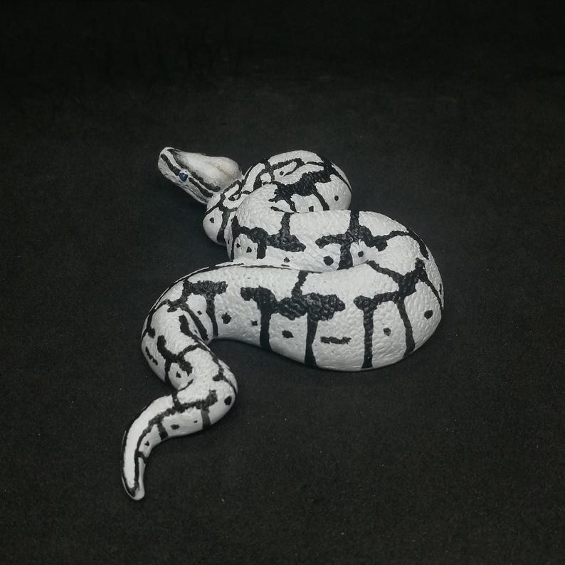 May include: A white and black snake figurine, coiled on a dark surface. The snake has a detailed pattern of black markings on a white base, with a realistic texture. The figurine is a decorative item.