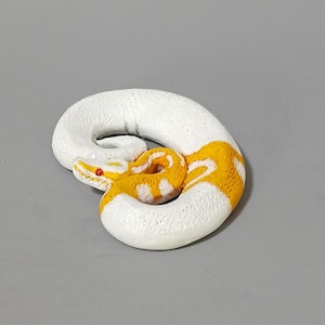 May include: A coiled, white snake figurine with yellow and white patterned markings. The snake has a red eye and is resting on a gray surface. The figurine is a decorative item.