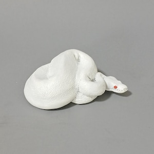 May include: A coiled white snake figurine with red eyes. The snake is sculpted with textured details, and its head is slightly raised. The background is a solid gray.