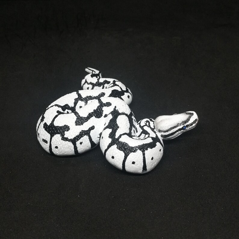 May include: A white and black decorative snake figurine. The snake is coiled and features a pattern of black markings on a white base. The head is detailed with black stripes and small blue accents. The figurine is set against a black background.