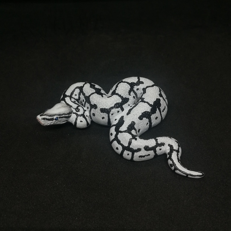 May include: A white and black patterned snake figurine. The snake is coiled and has a detailed design with black markings on a white base. The snake's head is visible, with small blue eyes.