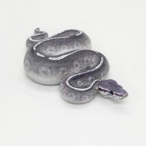 May include: A gray and white decorative snake figurine. The snake is coiled with a detailed head and body, featuring white stripes along its back. The figurine is set against a white background.