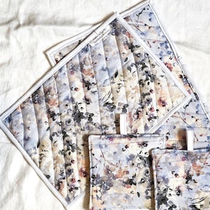 May include: Set of four quilted pot holders with a floral pattern in shades of gray, white, and brown. The pot holders have a white border and the text "Handmade by" is printed on one of the larger pot holders.