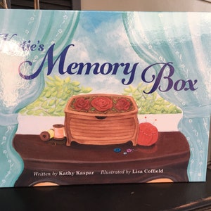 May include: A book titled "Katie's Memory Box" with a blue and white background. The cover features a wooden box with red roses on top, sitting on a table with sewing supplies. The book is written by Kathy Kaspar and illustrated by Lisa Coffield.