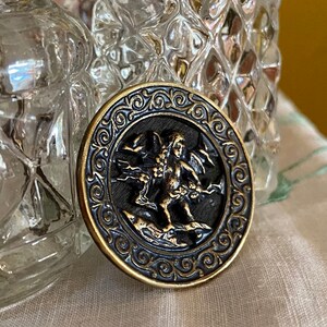 Pu&ograve; includere: Un bottone rotondo color oro con un centro nero che presenta una scultura a rilievo dettagliata di un cherubino alato che tiene un fiore. Il bottone &egrave; circondato da un bordo decorativo e ornato.
