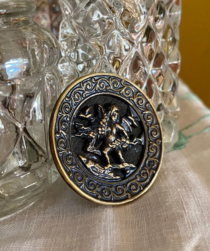Pu&ograve; includere: Un bottone rotondo color oro con un centro nero che presenta una scultura a rilievo dettagliata di un cherubino alato che tiene un fiore. Il bottone &egrave; circondato da un bordo decorativo e ornato.