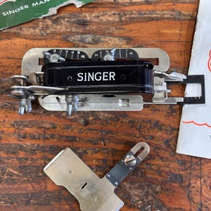 May include: Vintage Singer Manufacturing Company sewing machine attachment. The attachment is black and silver with the Singer logo. The image also shows a detached metal piece and a green and white box.