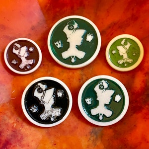 May include: Five round buttons featuring white silhouettes of a person wearing a hat and a dress. The buttons have different coloured backgrounds: green, brown, black, and two more green buttons.