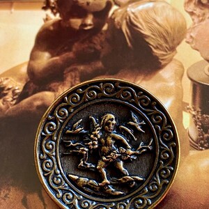 Pu&ograve; includere: Un bottone circolare di colore bronzo con un bordo ornato da volute. Al centro, un'immagine in rilievo di un cherubino alato che tiene un ramo con foglie e bacche.