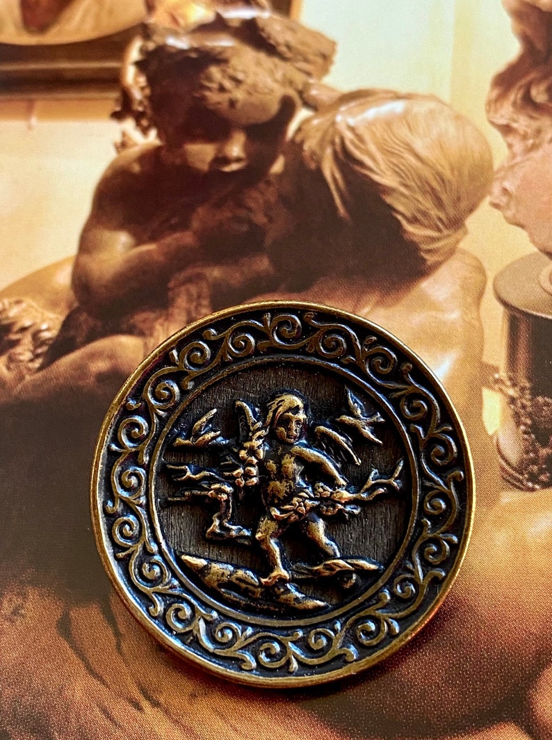 Pu&ograve; includere: Un bottone circolare di colore bronzo con un bordo ornato da volute. Al centro, un'immagine in rilievo di un cherubino alato che tiene un ramo con foglie e bacche.