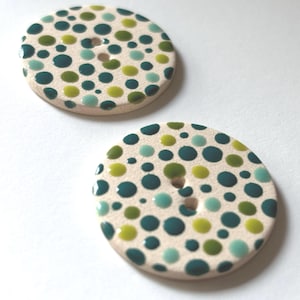 May include: Two round ceramic buttons with a white background and a polka dot pattern in shades of teal, green, and yellow.