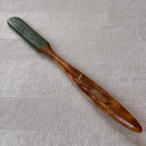 May include: A wooden butter spreader with a green top and a brown handle. The spreader is made of wood and has a smooth, polished finish. The handle is long and slender, and the top is rectangular.