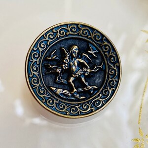 Pu&ograve; includere: Un bottone rotondo color oro con un designo blu e oro. Il bottone presenta una figura di cherubino con ali, che tiene un fiore e sta in piedi su uno scoglio. Il bottone &egrave; circondato da un bordo decorativo.