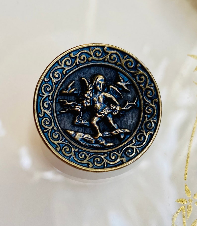Pu&ograve; includere: Un bottone rotondo color oro con un designo blu e oro. Il bottone presenta una figura di cherubino con ali, che tiene un fiore e sta in piedi su uno scoglio. Il bottone &egrave; circondato da un bordo decorativo.