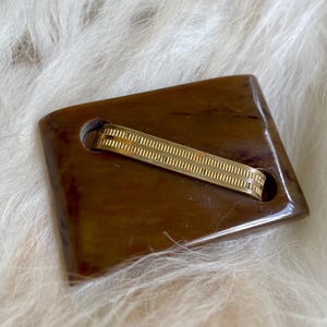 May include: A vintage brown rectangular brooch with a gold-colored metal band across the center. The brooch has a marbled appearance and rounded edges. The background is a soft, white, fluffy texture.
