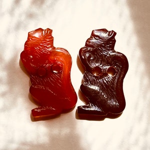 May include: Two vintage plastic buttons shaped like squirrels. The button on the left is a reddish-brown colour and the button on the right is a dark brown colour.