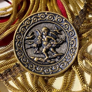 Pu&ograve; includere: Un bottone rotondo color oro con un designo dettagliato in nero e oro. Il centro del bottone presenta un'immagine in rilievo di un cherubino alato che tiene un fiore.
