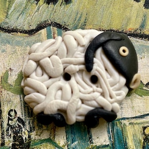 May include: A white and black sheep button made of polymer clay. The sheep is curled up with its head tucked in. The sheep has black eyes and a black nose.