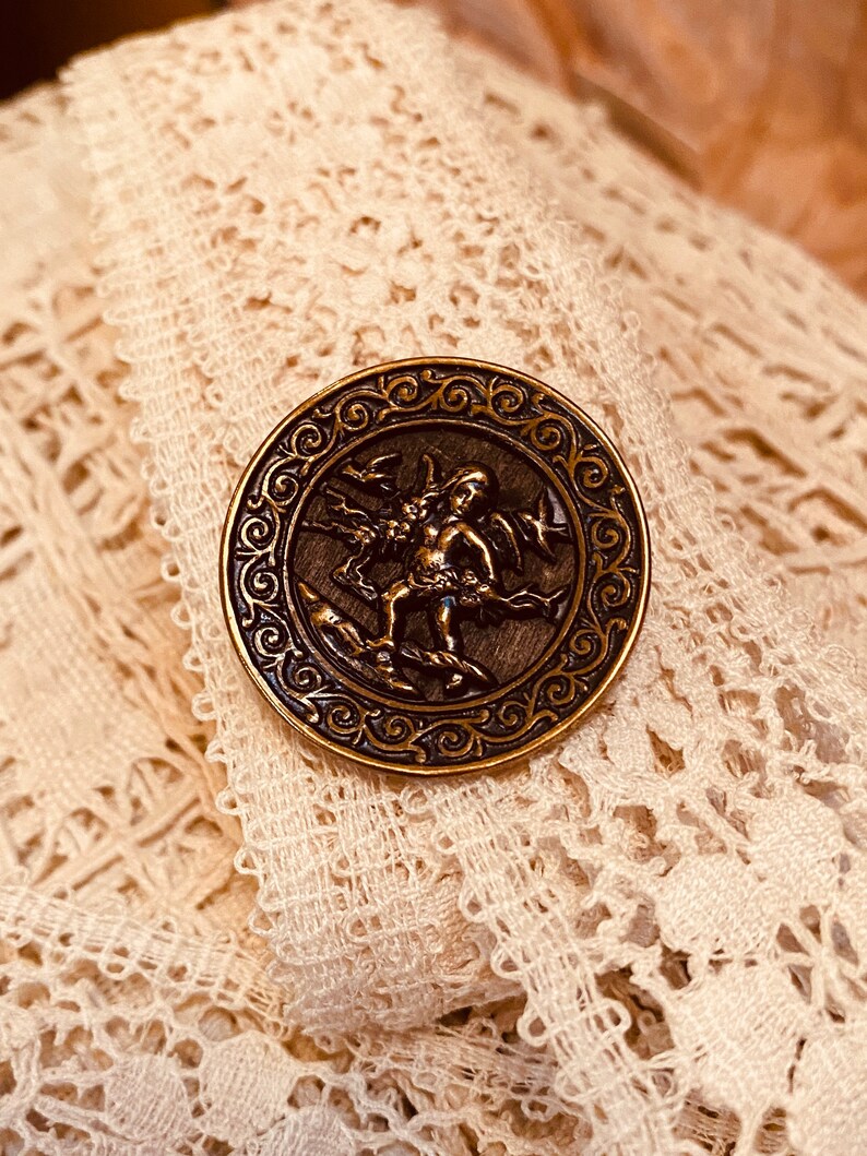Pu&ograve; includere: Un bottone in metallo dorato con un design ornamentale che presenta una figura di cherubino con ali e un uccello. Il bottone &egrave; posto su uno sfondo di pizzo bianco.
