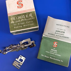 May include: A vintage Singer zig-zag attachment for sewing machines, including a white and green cardboard box, a metal attachment mechanism, a separate metal presser foot plate, and a printed instruction booklet for the Singer adjustable attachment.