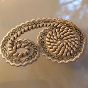 May include: A white and silver embroidered design with a paisley shape. The design is made of a braided rope-like material and has a spiral pattern.