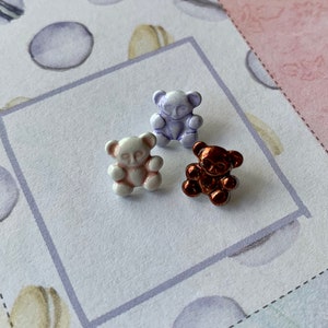 May include: Three small teddy bear buttons in different colors: white, light purple, and brown. The buttons are on a white background.