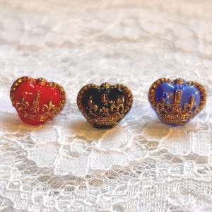 May include: Three crown-shaped buttons featuring a metallic gold-tone finish with intricate detailing. The buttons are coloured in glossy red, black, and blue, each displaying a raised fleur-de-lis pattern and a textured base rim design.