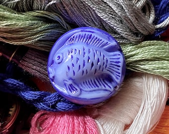 May include: A blue glass button with a raised design of a fish. The button is surrounded by colorful embroidery floss.