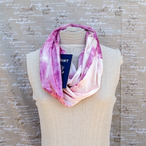 May include: A pink and white tie-dye scarf is draped around a mannequin's neck. A blue passport is tucked into the scarf. The background is a beige fabric with a repeating pattern of text.