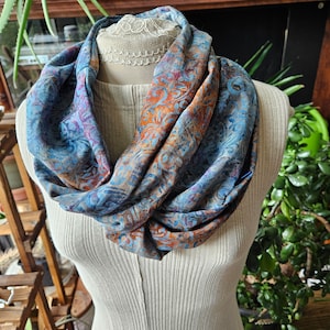 May include: A blue, orange, and purple patterned infinity scarf displayed on a mannequin. The scarf has a swirling design with floral and paisley elements. The mannequin is wearing a cream-colored ribbed top. The background includes plants and wooden shelves.