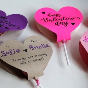 May include: Three heart-shaped Valentine's Day cards with lollipop sticks. The cards are decorated with pink, purple, and brown paper. The text on the cards reads "happy Valentine's day", "To: Sofia", and "From: Amelie Thanks for making life so sweet!"