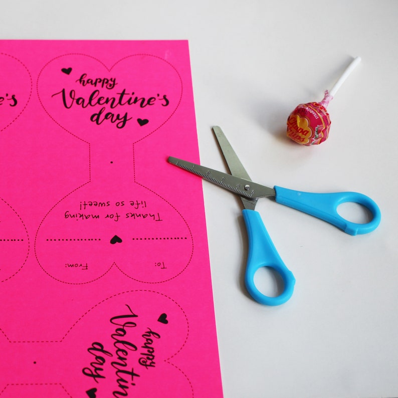 May include: A pink cardstock sheet with two heart-shaped Valentine's Day cards printed on it. The cards say "Happy Valentine's Day" and have a space for "To:" and "From:". There are also scissors and a lollipop on the table.