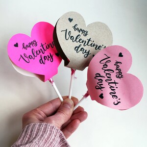 May include: Three heart-shaped lollipops with sticks. The lollipops are pink and brown with the text "happy valentine's day" written in black.