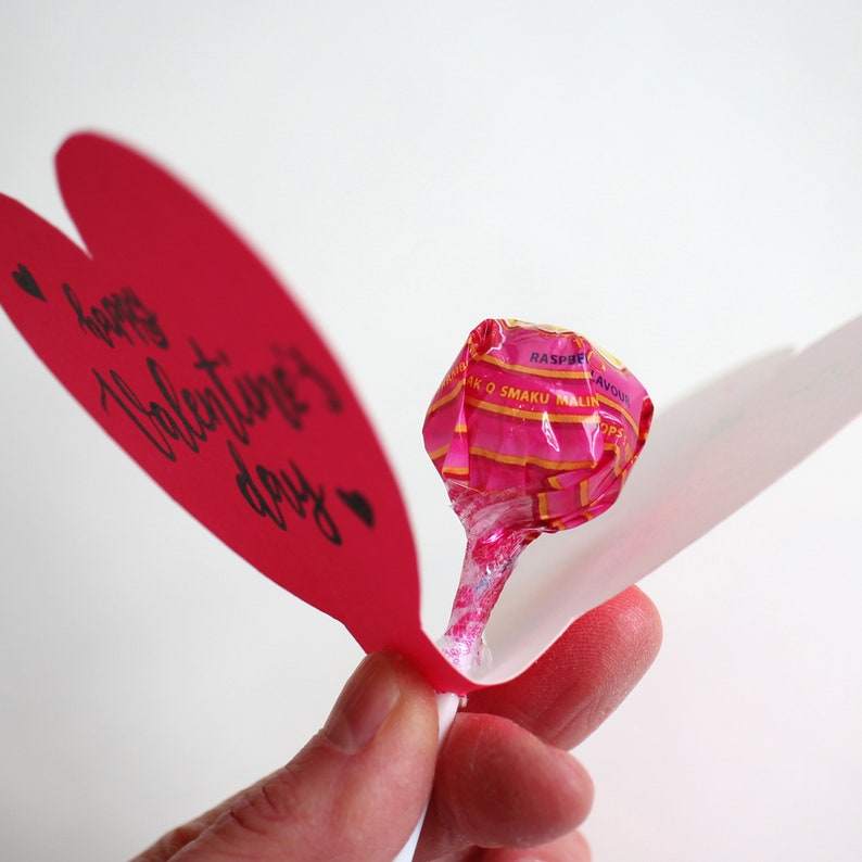 May include: A red paper heart with the text "Happy Valentine's Day" and two small hearts. A pink lollipop with a white stick is sticking out of the heart.