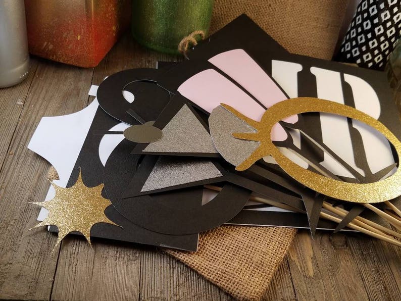 May include: A collection of photo booth props in various shapes and colors. Includes a gold glitter starburst, a gold glitter frame, and black cutouts spelling out "WED". Some props have wooden sticks attached.