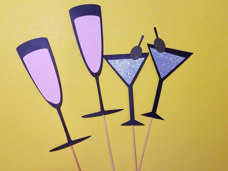 May include: Four cocktail-themed photo booth props against a yellow background. Two champagne flutes and two martini glasses are depicted. The flutes have pink interiors, while the martini glasses have silver glitter interiors and olive garnishes.