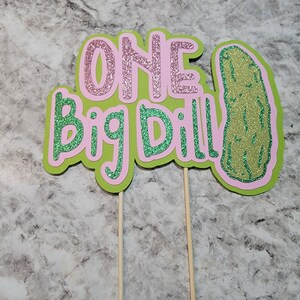 Op de afbeelding: Twee groene en roze glittertaarttoppers met de tekst "ONE Big Dill!" en een groene glitteraugurk.