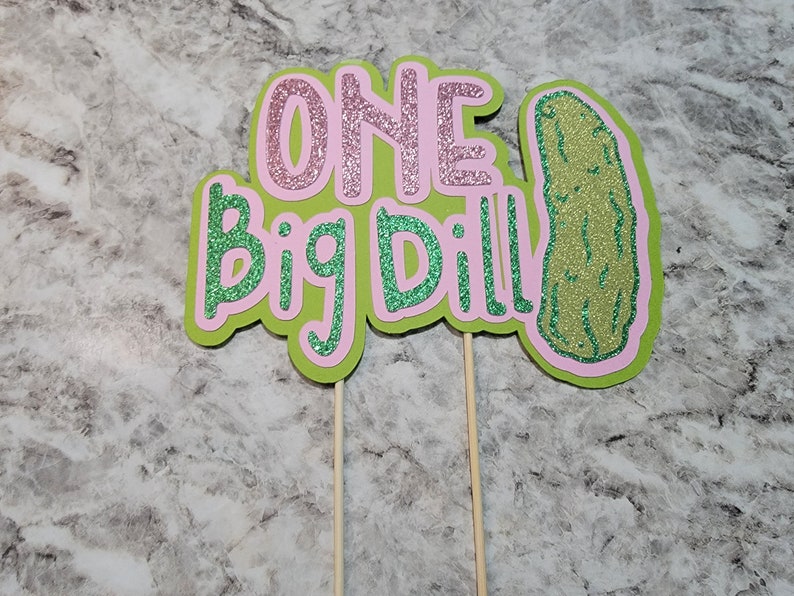 Op de afbeelding: Twee groene en roze glittertaarttoppers met de tekst "ONE Big Dill!" en een groene glitteraugurk.