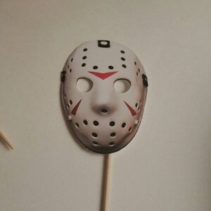 May include: White hockey mask with black eye holes and red accents on a wooden stick. The mask is a popular Halloween decoration.