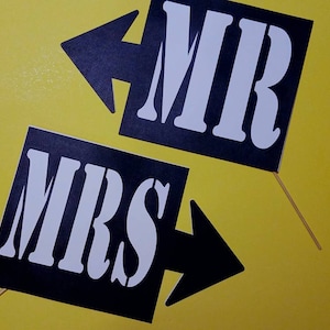 May include: Two black signs with white text that read "MR" and "MRS" with arrow designs. The signs are on wooden sticks and set against a yellow background. Ideal for wedding or engagement photos.
